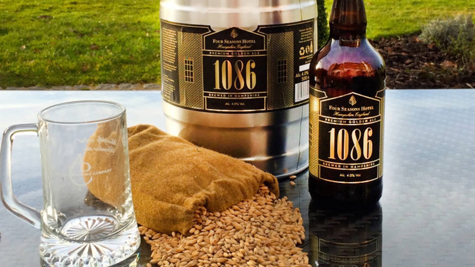 Bottle, keg of Four Seasons 1086 craft ale on garden table with barley spilling from burlap bag, glass