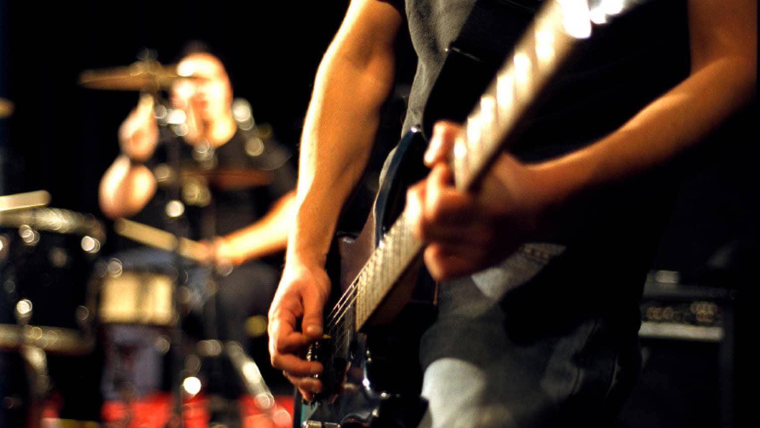 Close-up of musician's arms and hands playing an electric guitar with drumset in the background.