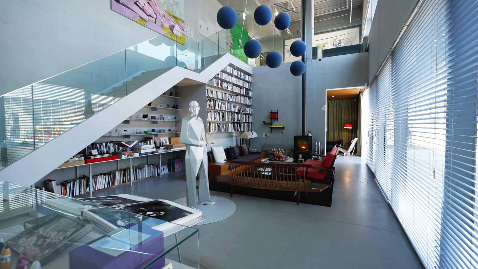 A spacious modern room with a glass staircase, a wall of bookshelves, seating area and several contemporary art pieces 