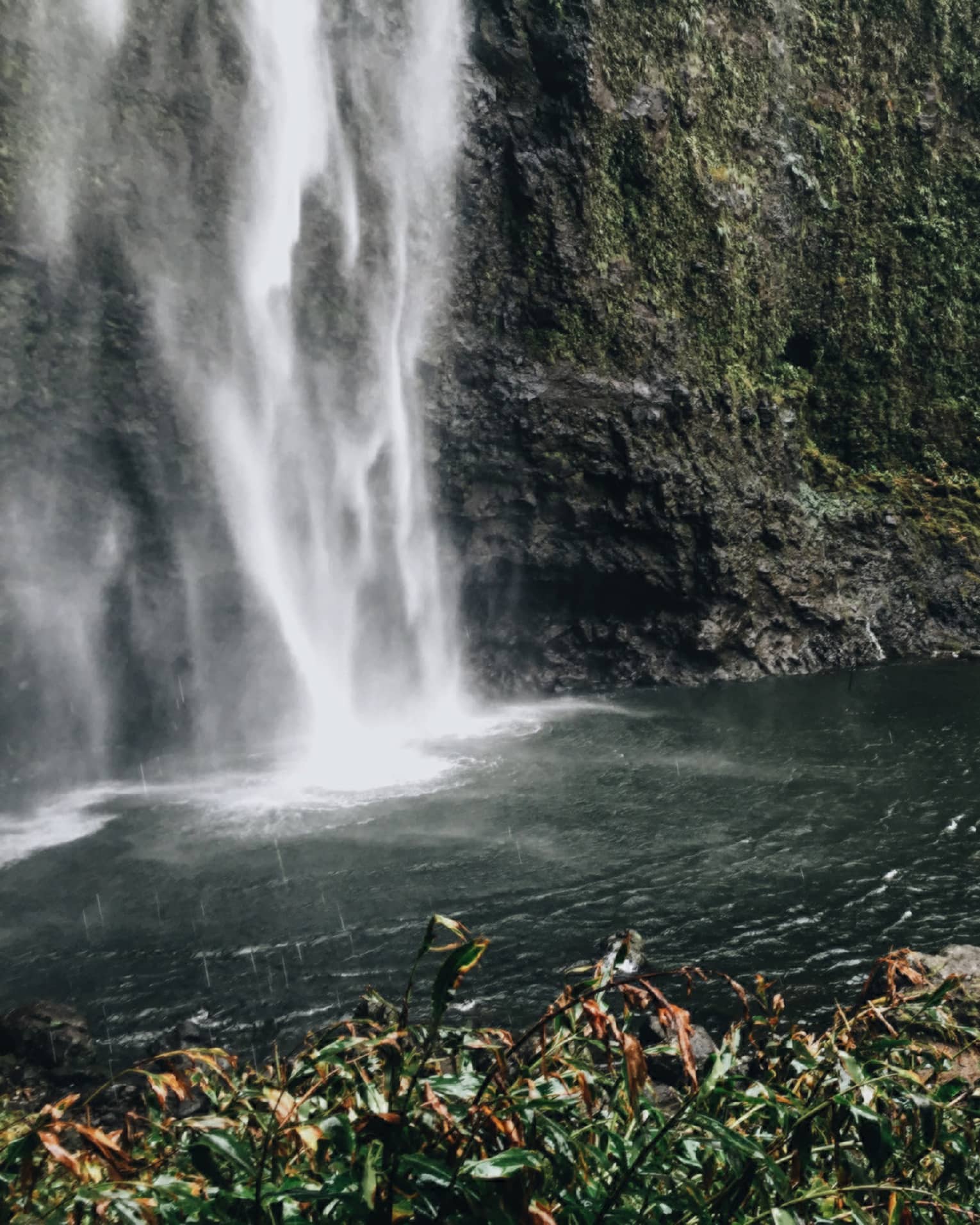 Back of woman hiking over black rocks under tall waterfall, small pond