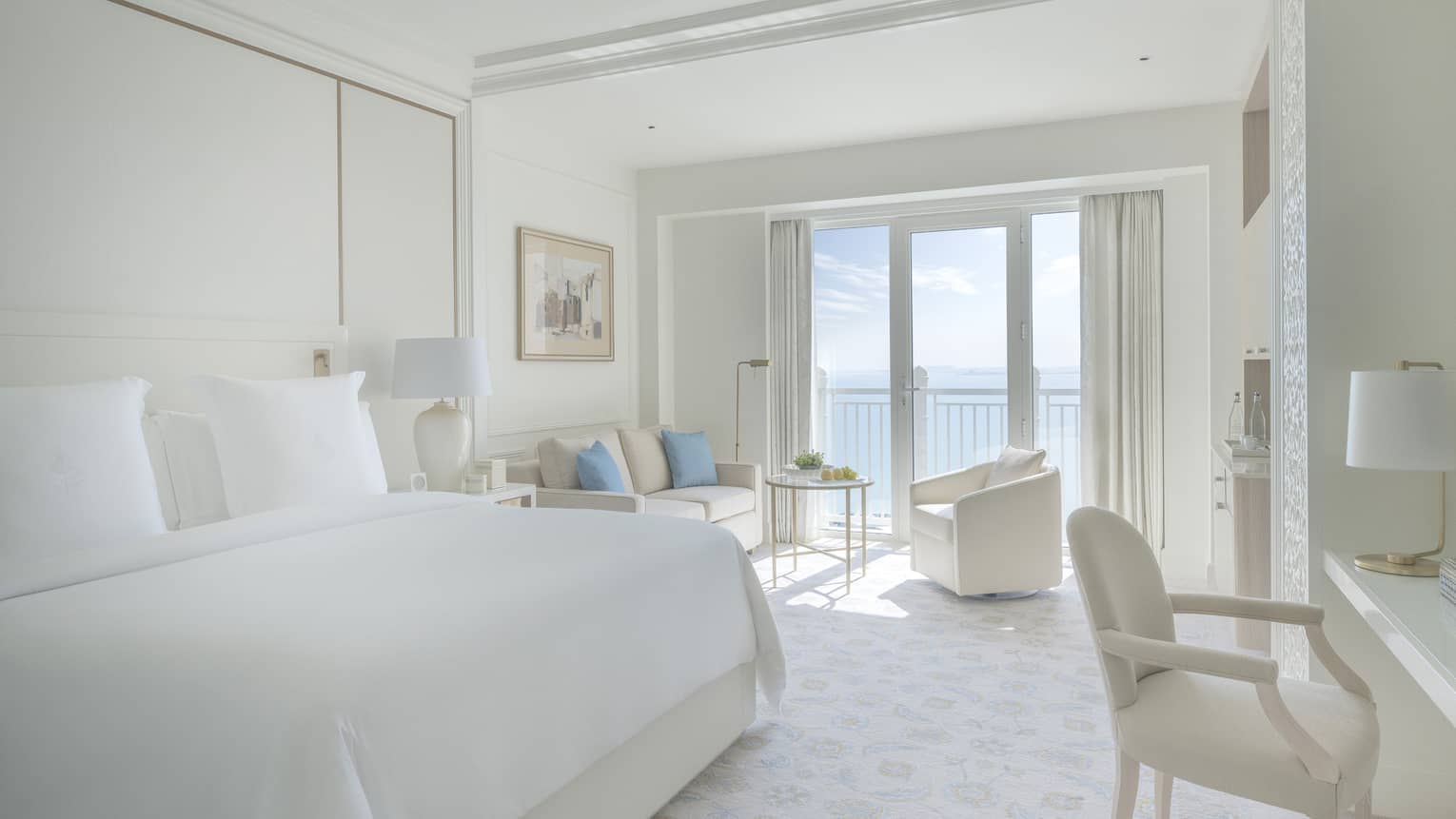 Bedroom with white bed, white walls and a white sofa and arm chair, with balcony offering sea view