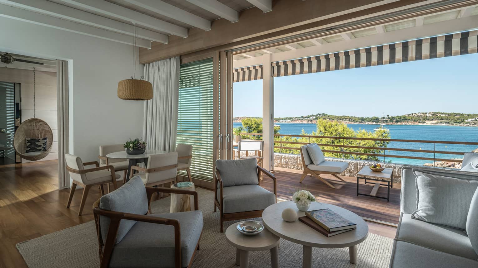 Bungalow Suite living room with grey arm chairs and nesting coffee table, dining table with four chairs, doorway to bedroom, balcony with ocean view