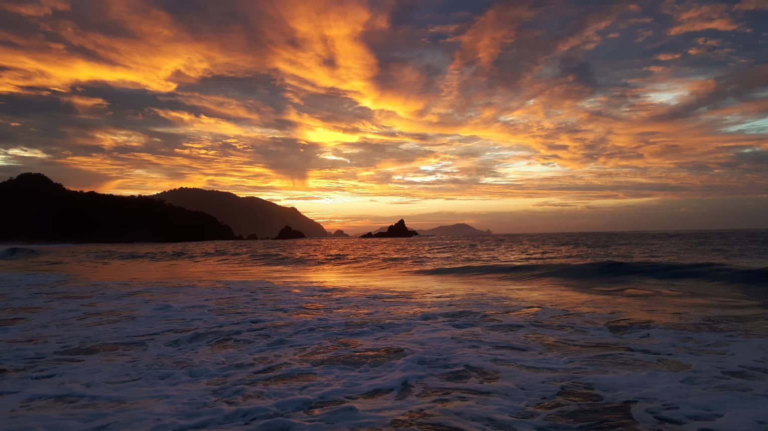 Waves foam at the shore in the foreground as darkness falls, distant hills and iridescent clouds glowing orange and gold. 