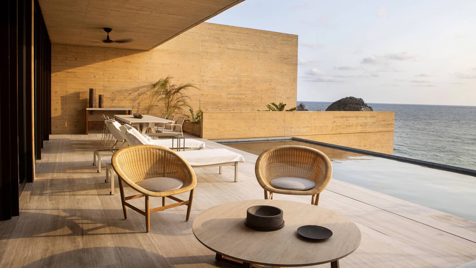 A modern oceanfront terrace featuring wicker chairs, a dining area and lounge chairs beside an infinity pool, all bathed in warm sunset light with a view of the sea
