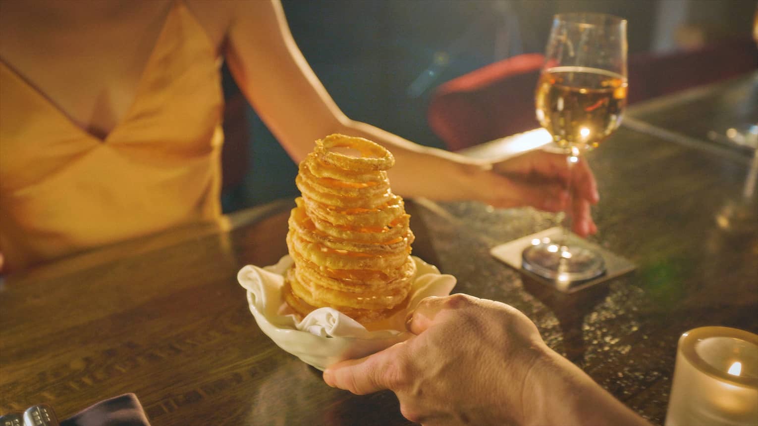 Stack of crispy onion rings on a small round white plate is being placed on a wooden table in front of a guest holding a glass of white wine