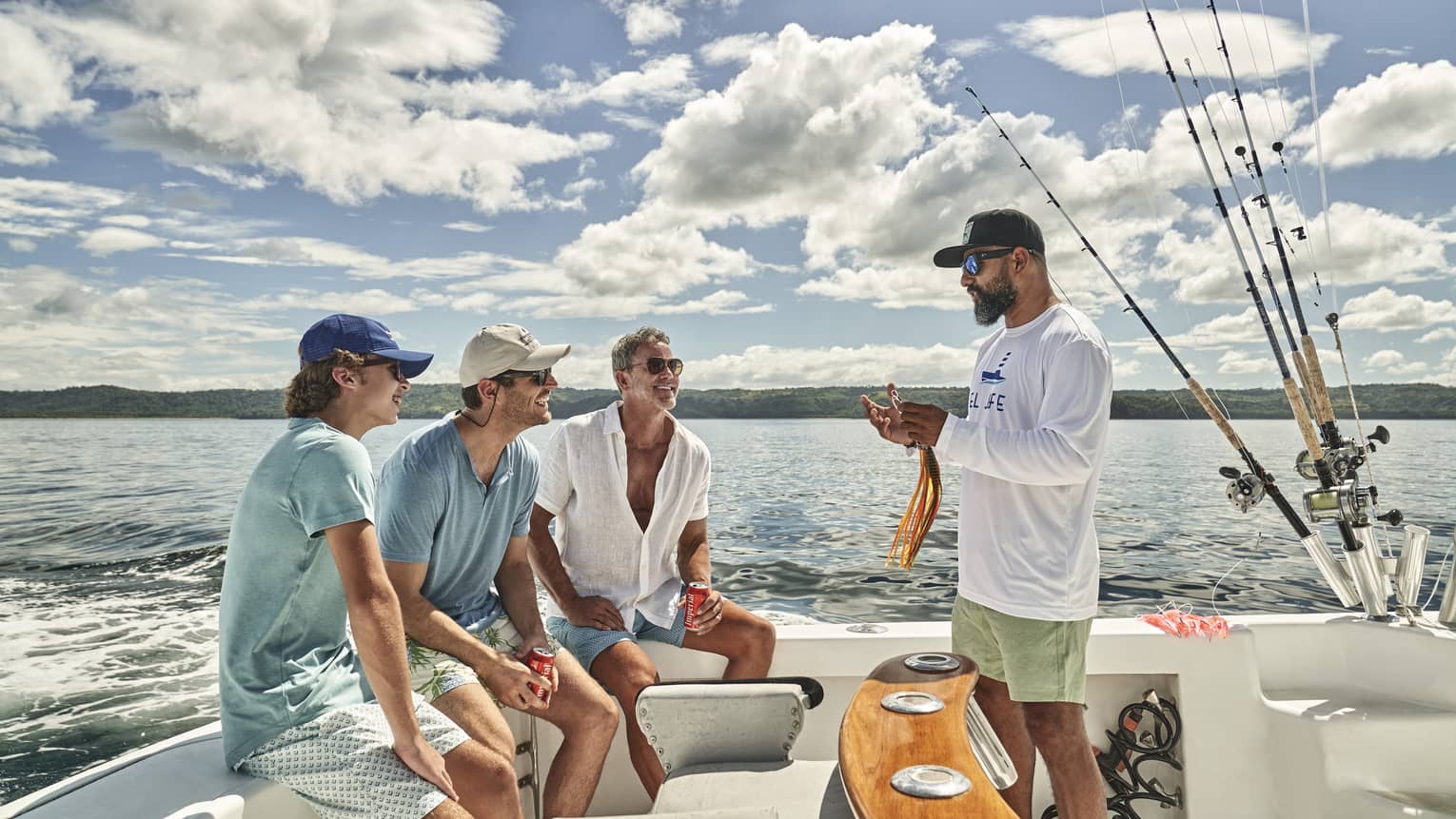 Aboard a fishing boat, a person presents a large colourful lure to a smiling group of three guests seated on the stern.