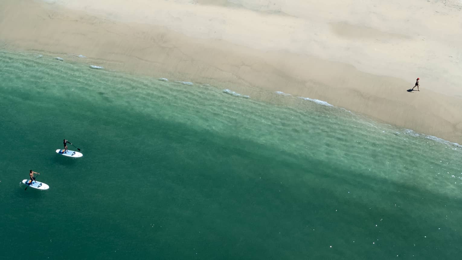 Aerial view of blue-green water lapping up on a beach, with two stand-up paddleboarders in the water and one person walking along the sand