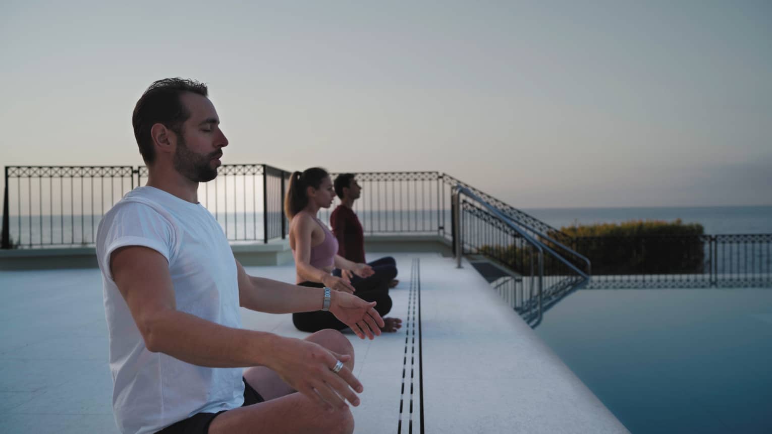 Under a grey sky, three guests sit cross-legged with eyes closed beside a calm pool bordered by greenery and the vast ocean.