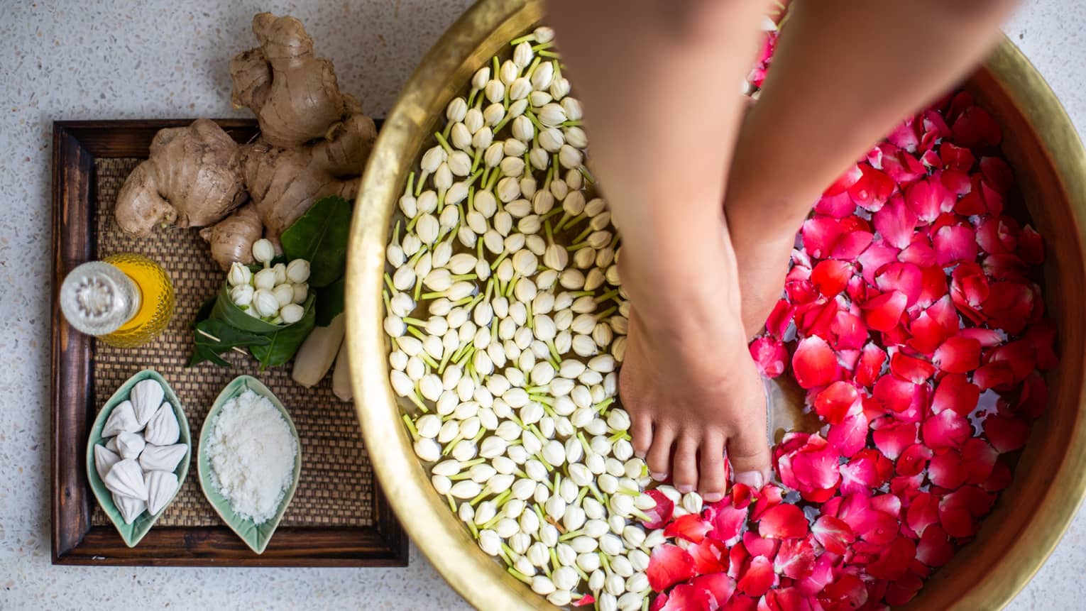 Woman's feet soaking in bowl of water with pink flower petals and white flower buds, tray of ginger and makrut lime leaves next to bowl