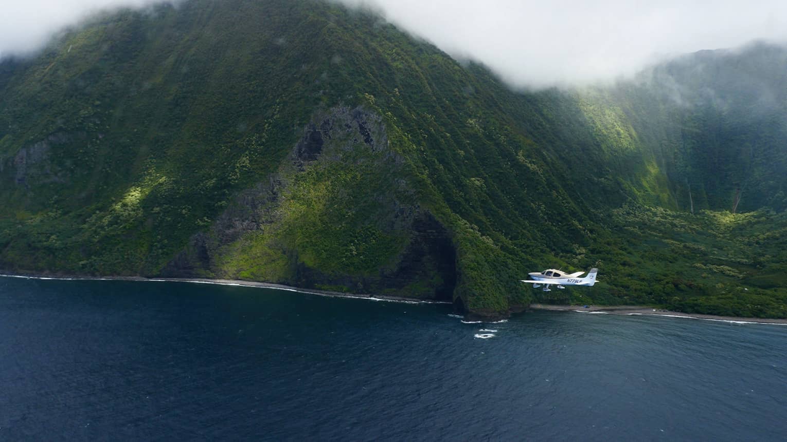 Small white airplane flies by large green mountain on Hawaii, Lanai coast