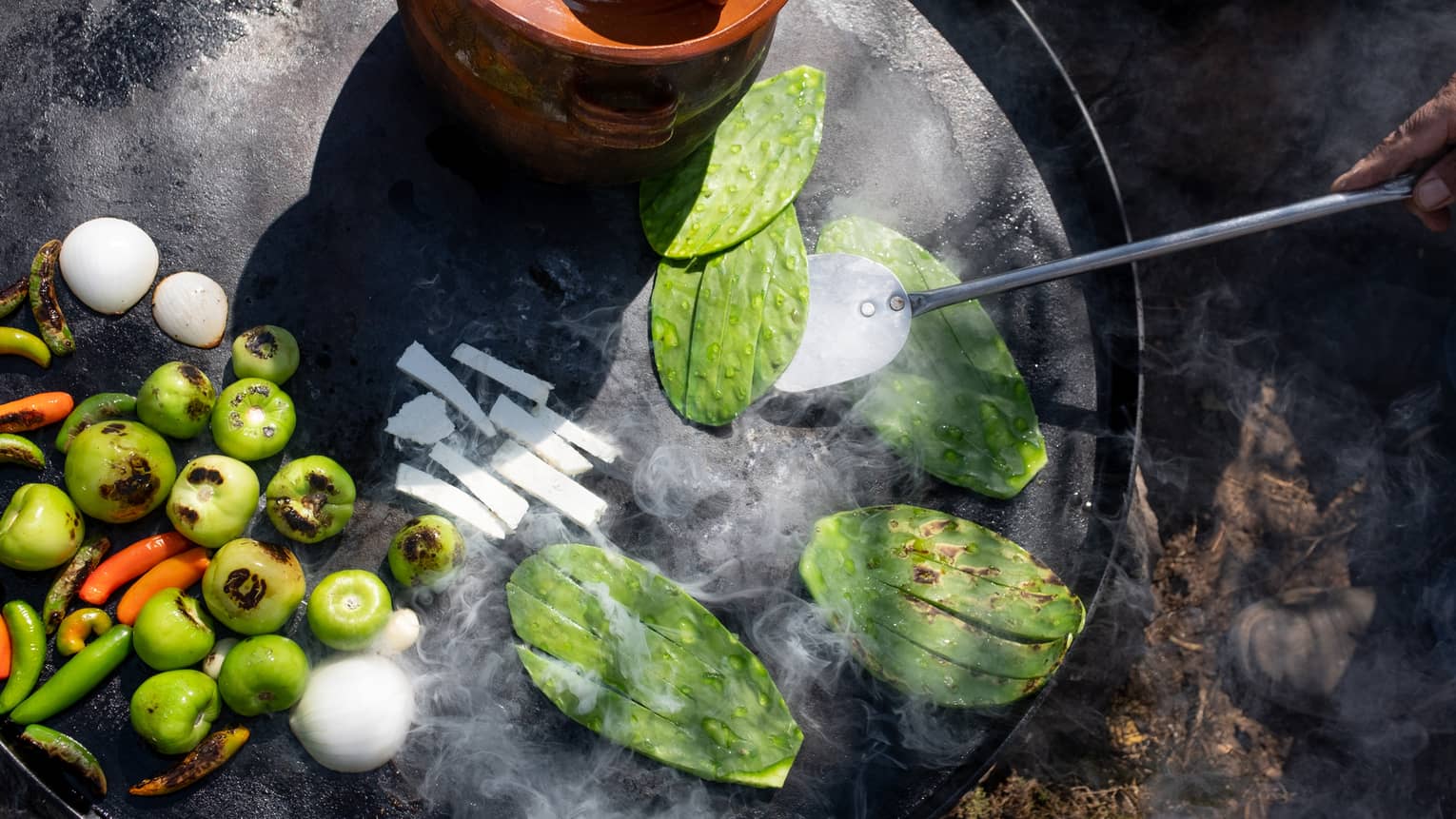 A charred cactus pad being flipped on a large cast-iron skillet beside frying tomatillos, white onions, and red hot peppers.