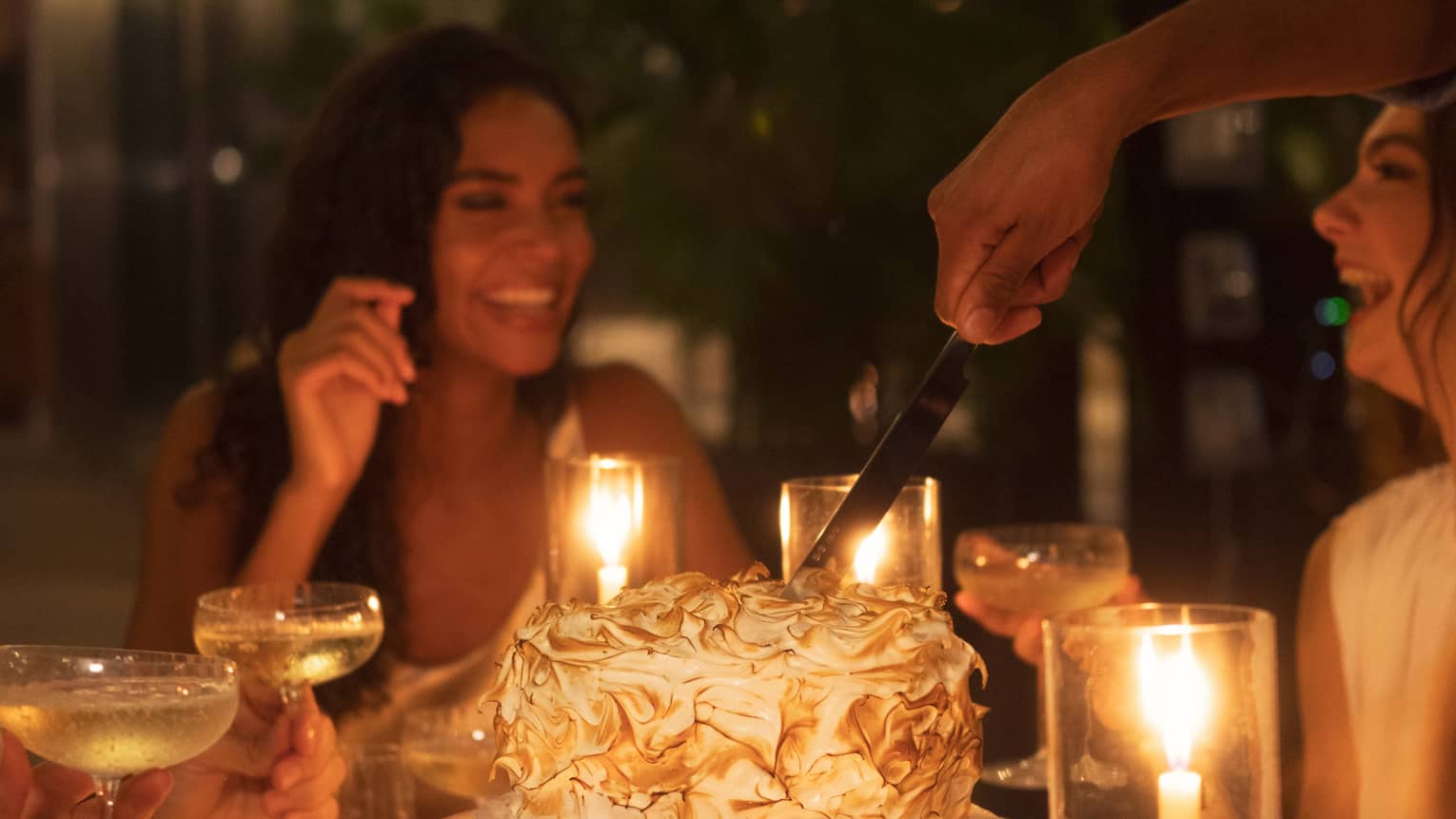 Two women at candle-lit dining table laugh while person cuts a meringue cake