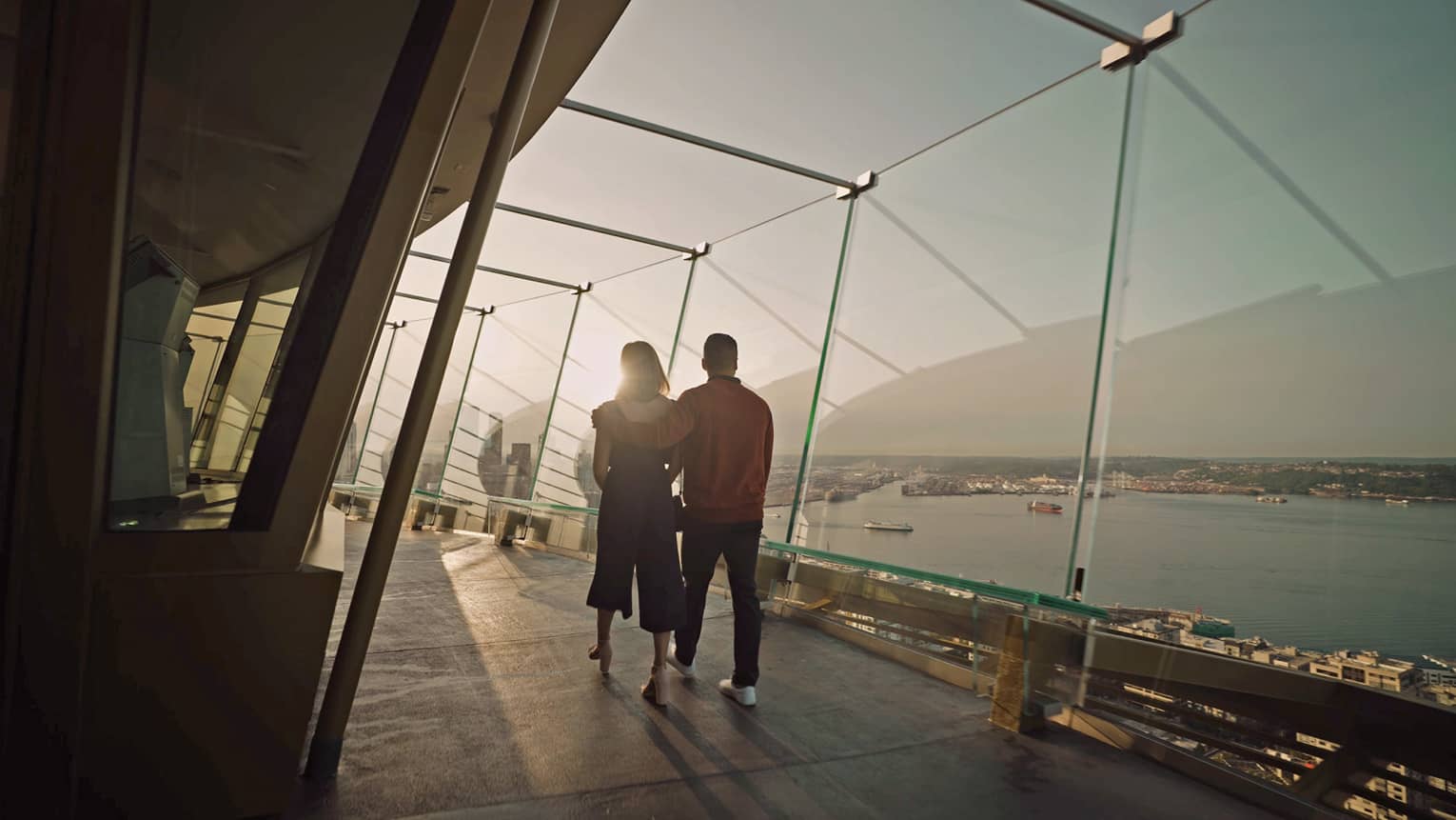 Rear view of couple walking along glass-walled walkway overlooking water