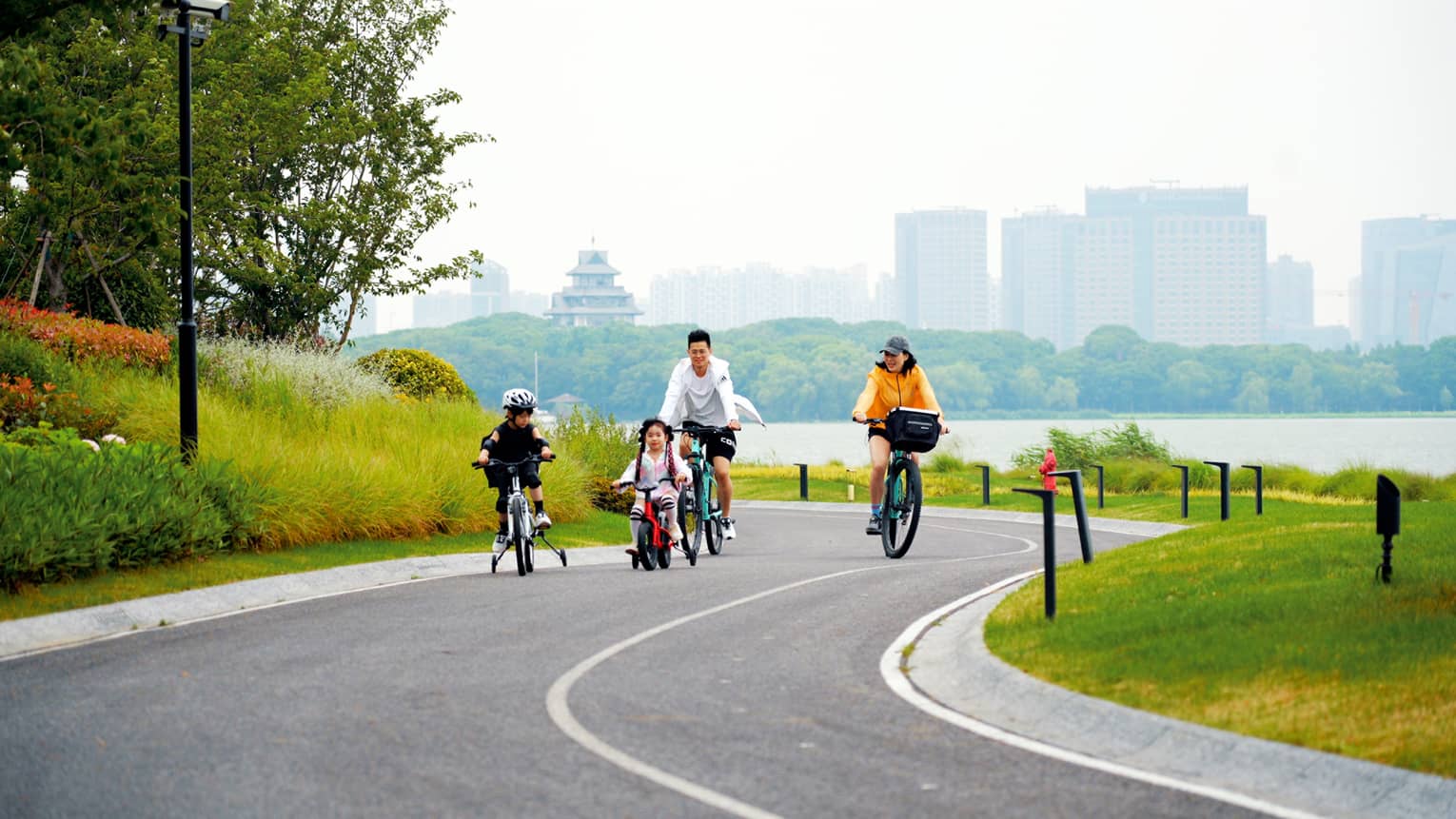 A family cycles uphill on a winding road bordered by bright grass and foliage, a hazy lake, forest and cityscape behind them.