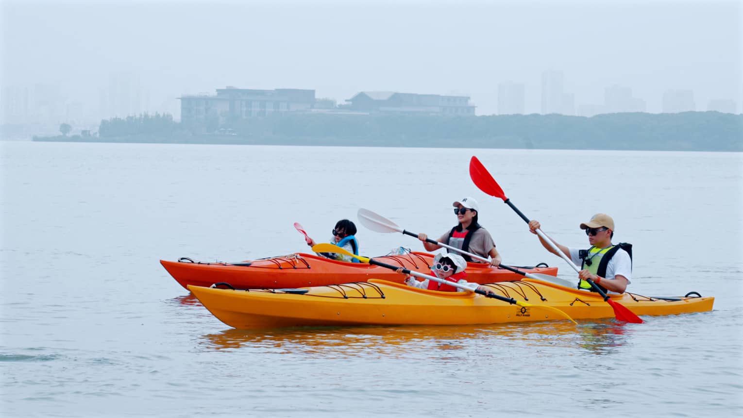 A family of four glides along serene waters, two people per kayak, distant terrain and buildings blurred by misty fog beyond.