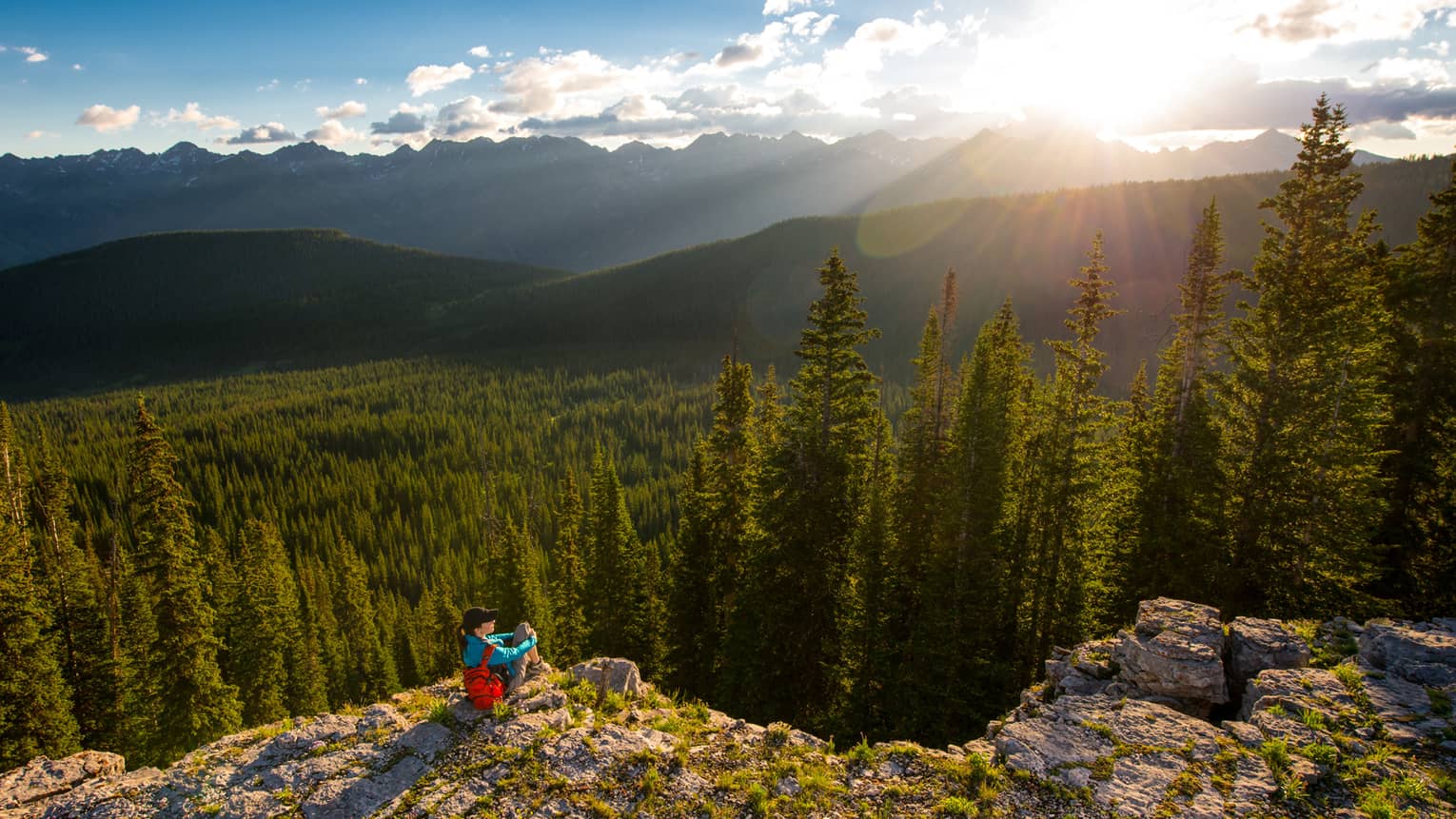 Person sits at edge of cliff overlooking trees, mountains as sun starts to set