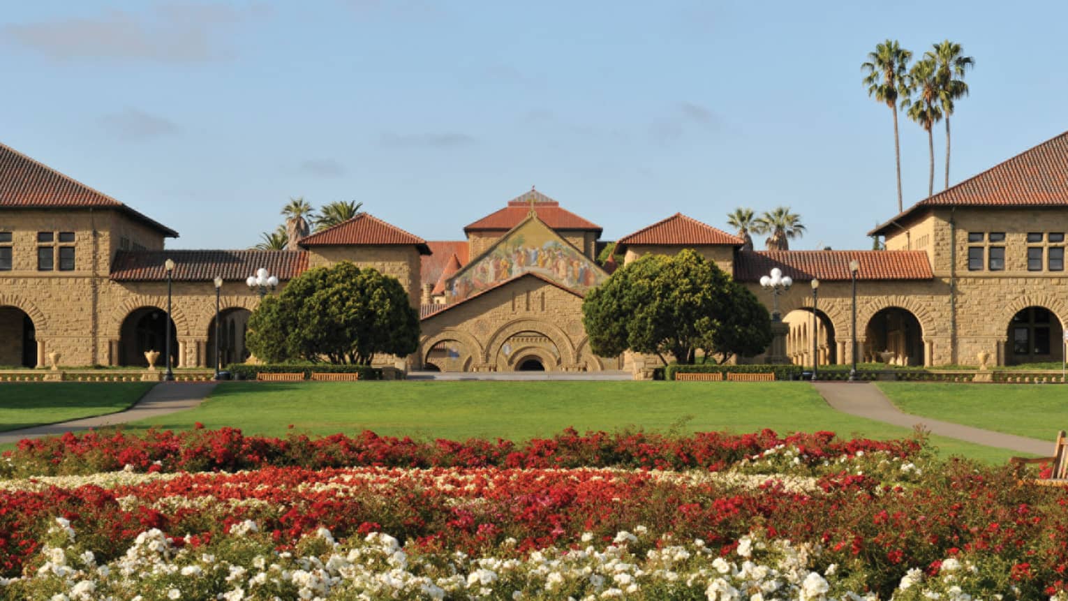 View across lawn and garden with red and white flowers to brick building, palm trees