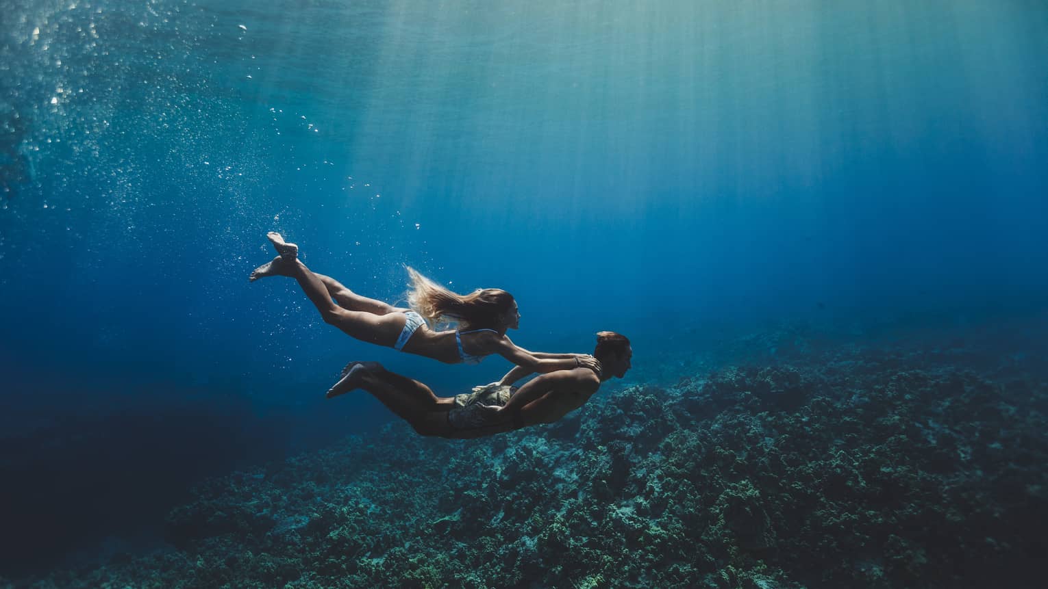 A man and woman swim underwater in the ocean over a coral reef. 