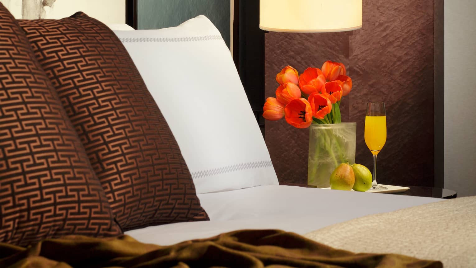 Deluxe Room close-up of white and brown pillows on bed with red tulips and mimosa glass on bedside table
