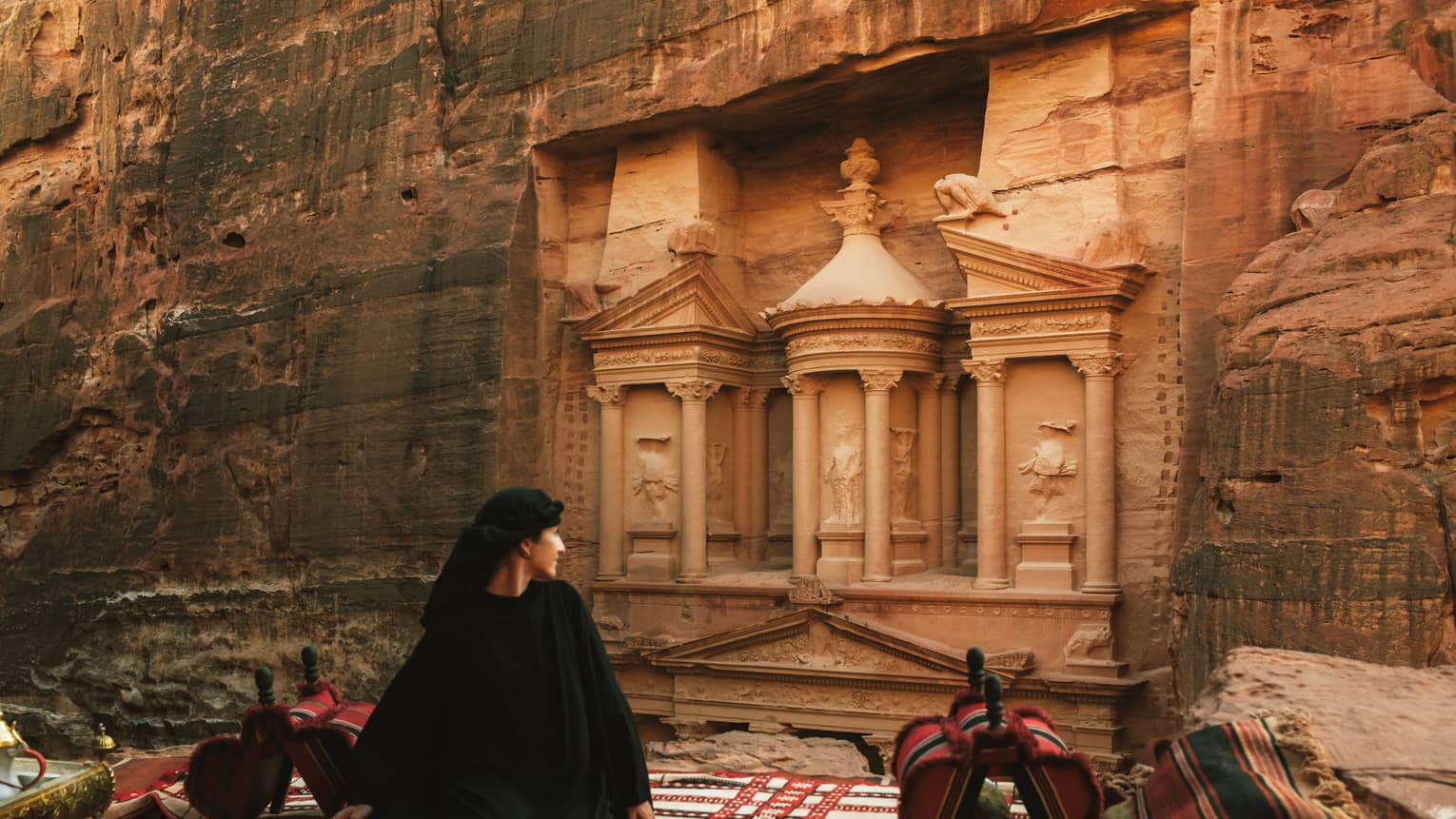 A person stands looking at buildings carved into a red rock cliffside.