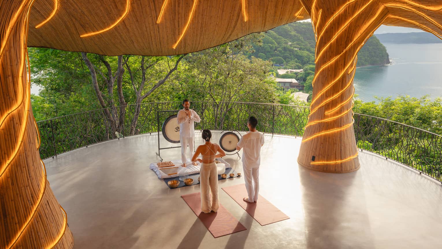 Two people stand on yoga mats facing an intructor who is standing in front of two gongs and surrounded by singing bowls, all beneath an organic-style pavillion lit with string lights and overlooking the ocean