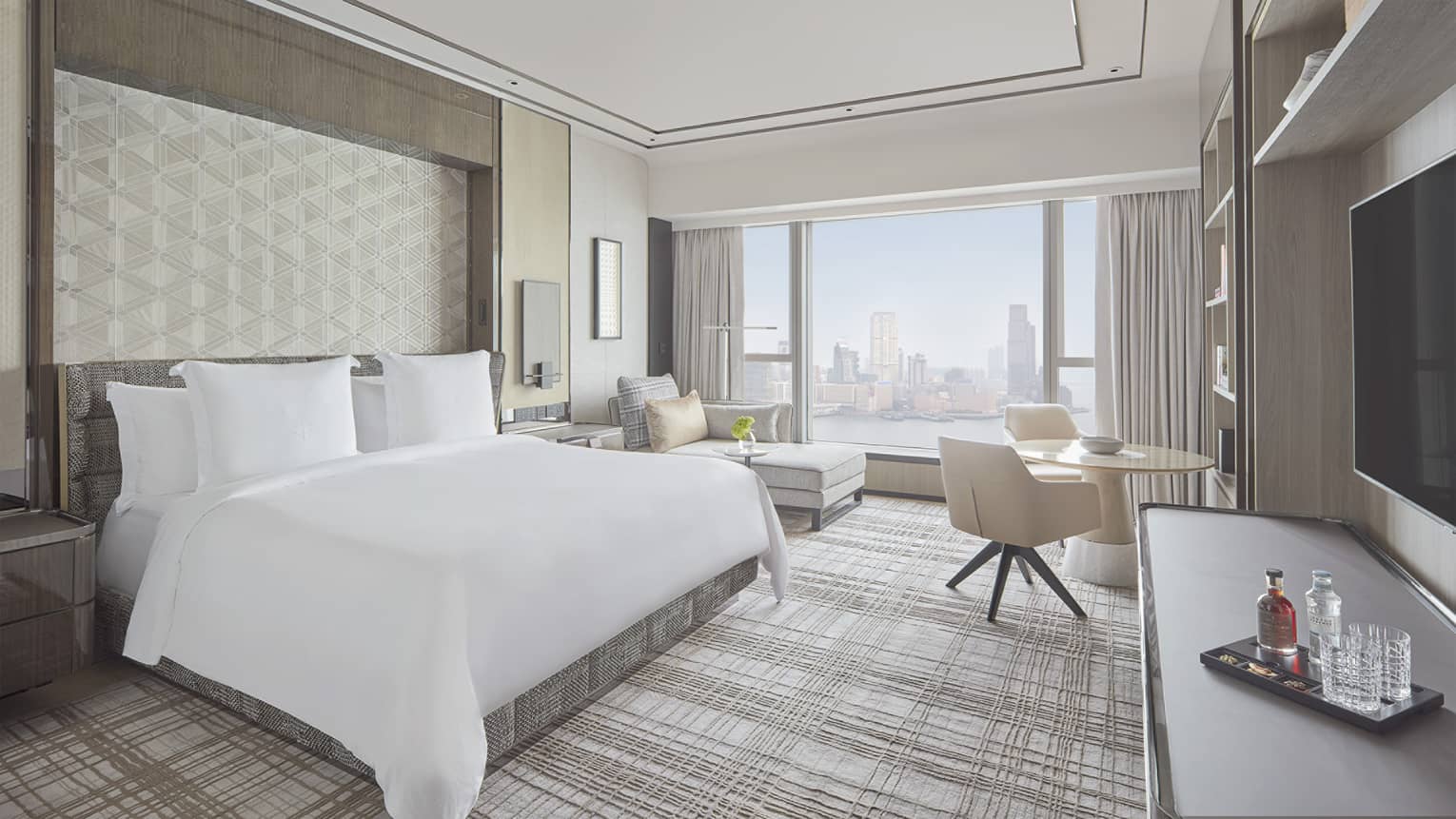 Hotel bedroom with king bed, TV, cocktail tray, round table with chair, chaise and window overlooking Hong Kong