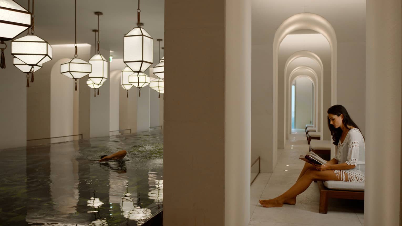 Woman sits reading a book on a lounge chair next to an indoor pool with multiple lanterns hanging over the water
