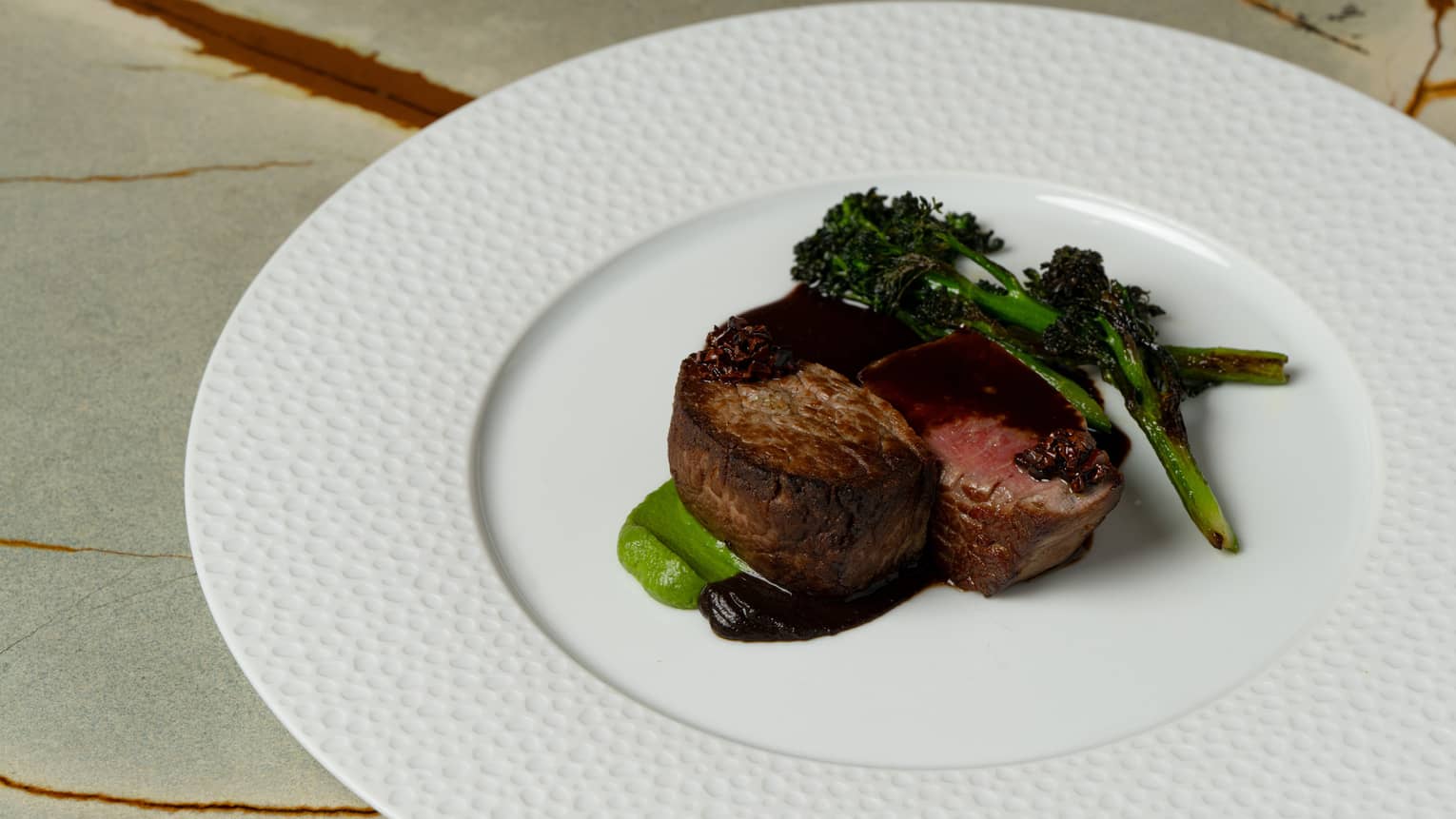 A round white plate holds pieces of cooked beef seated in a dollop of sauce, with cooked green vegetables on the side.