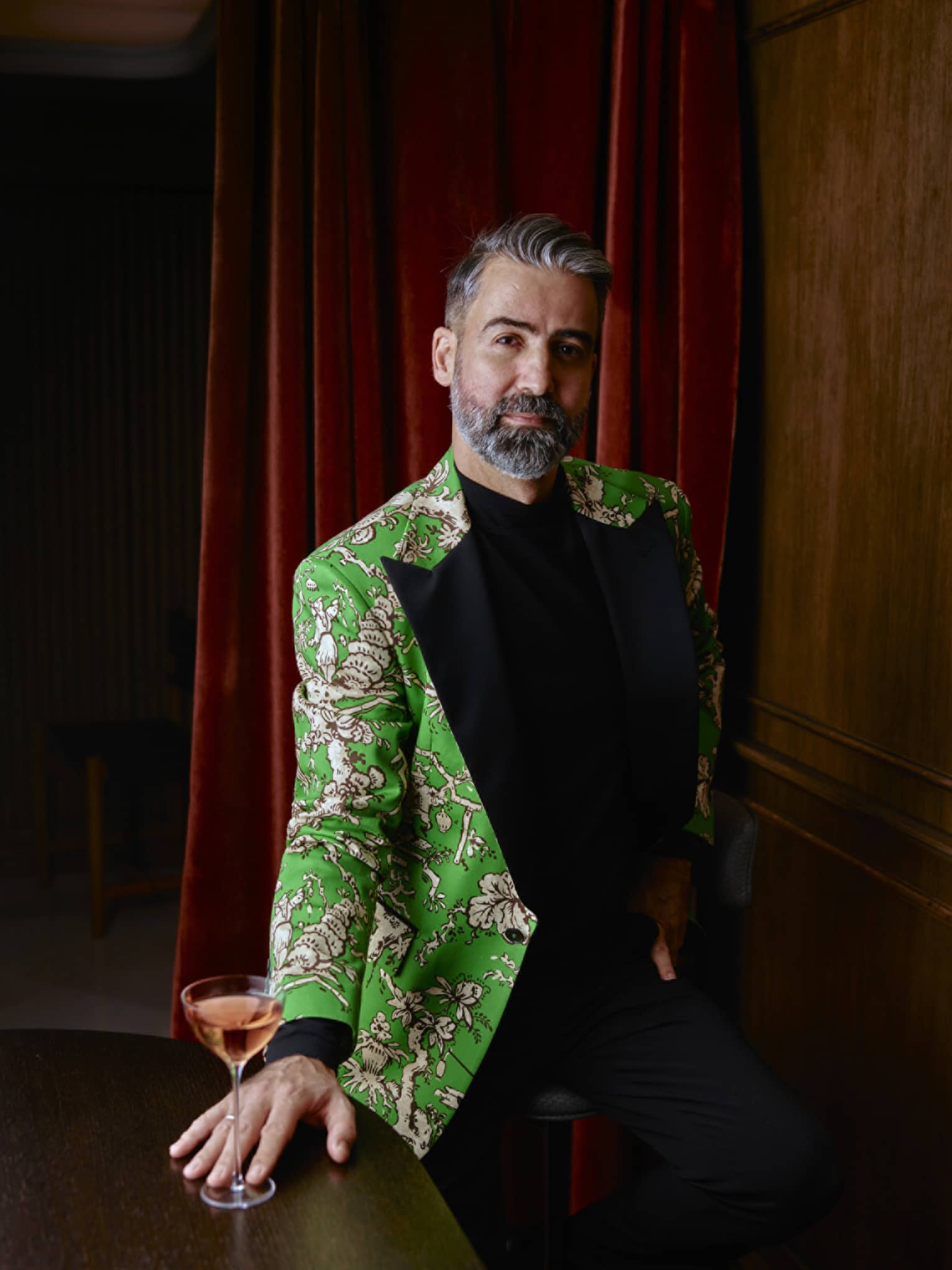A man in a green jacket with a floral print stands in front of a red curtain with a pink martini.