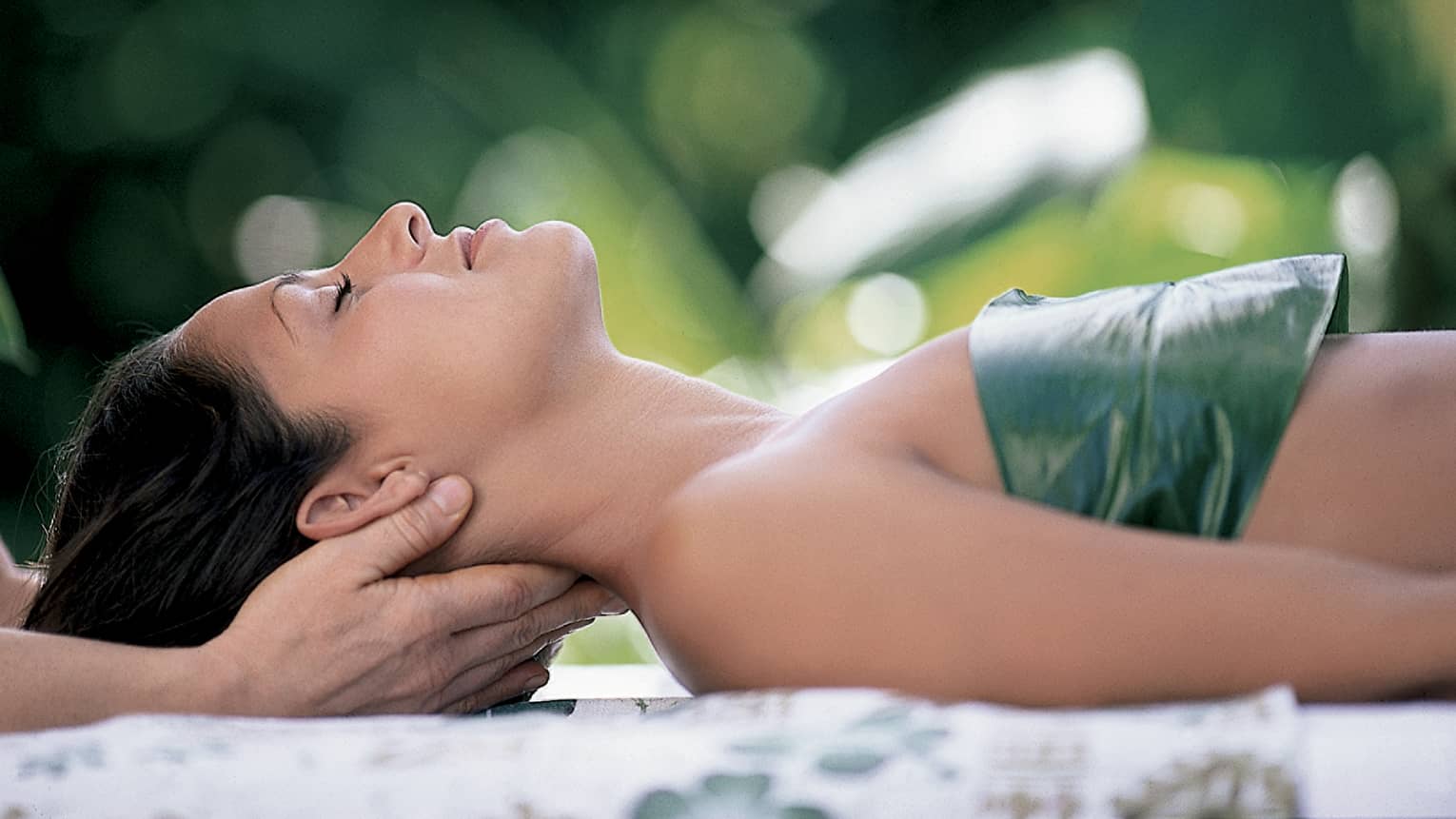 Woman with eyes closed lays on back on table with leaf over chest, hands massage her neck