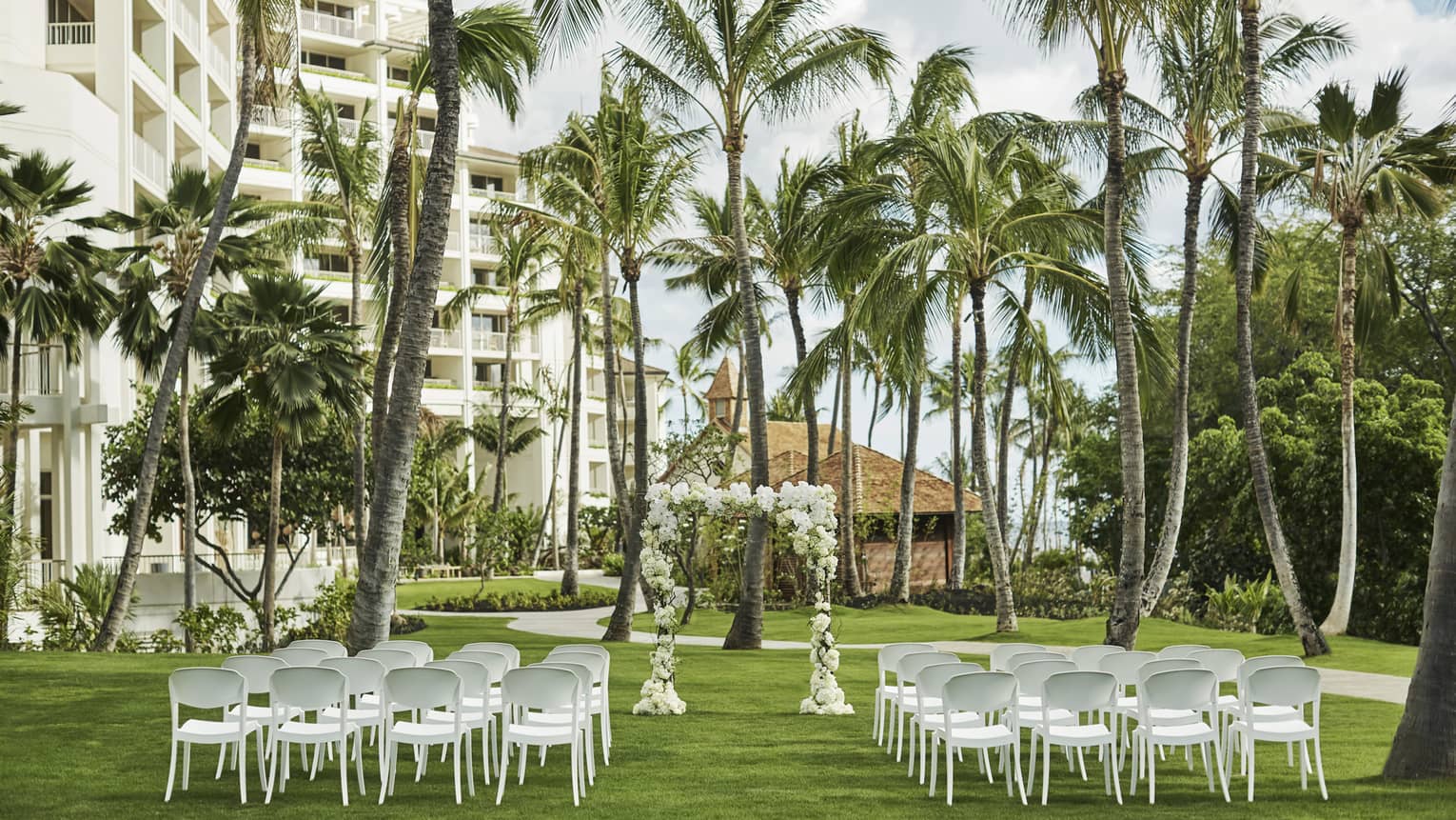 A picturesque outdoor wedding venue with elegant floral arrangements, white chairs on a lush green lawn, beneath palm trees