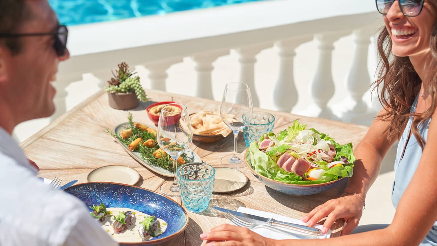 Couple smiling, dining on salad niçoise and other dishes beside white railing
