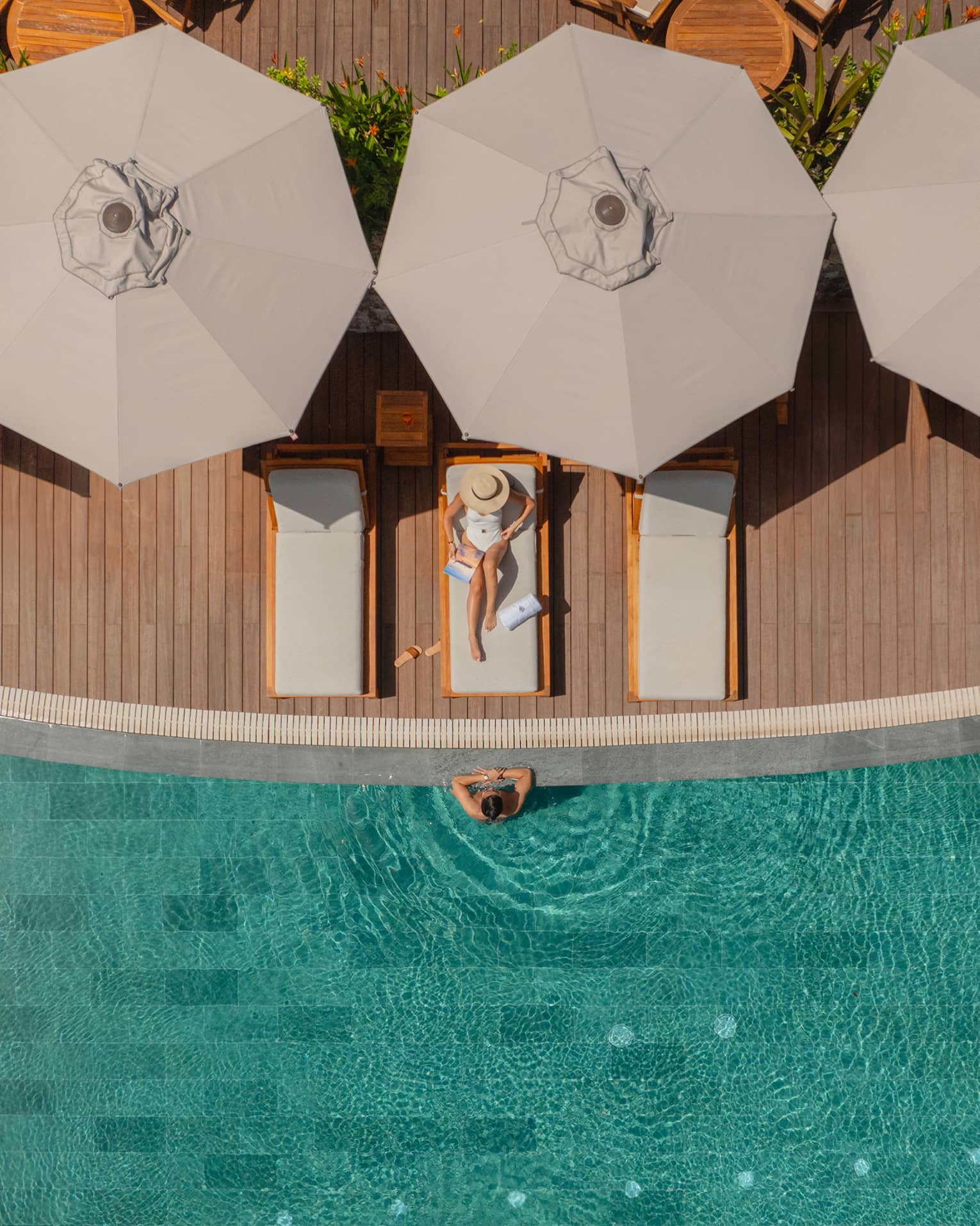 Aerial view of woman wearing white bathing suit relaxing on a lounge chair next to a pool with a man standing in the water with his arms on the ledge