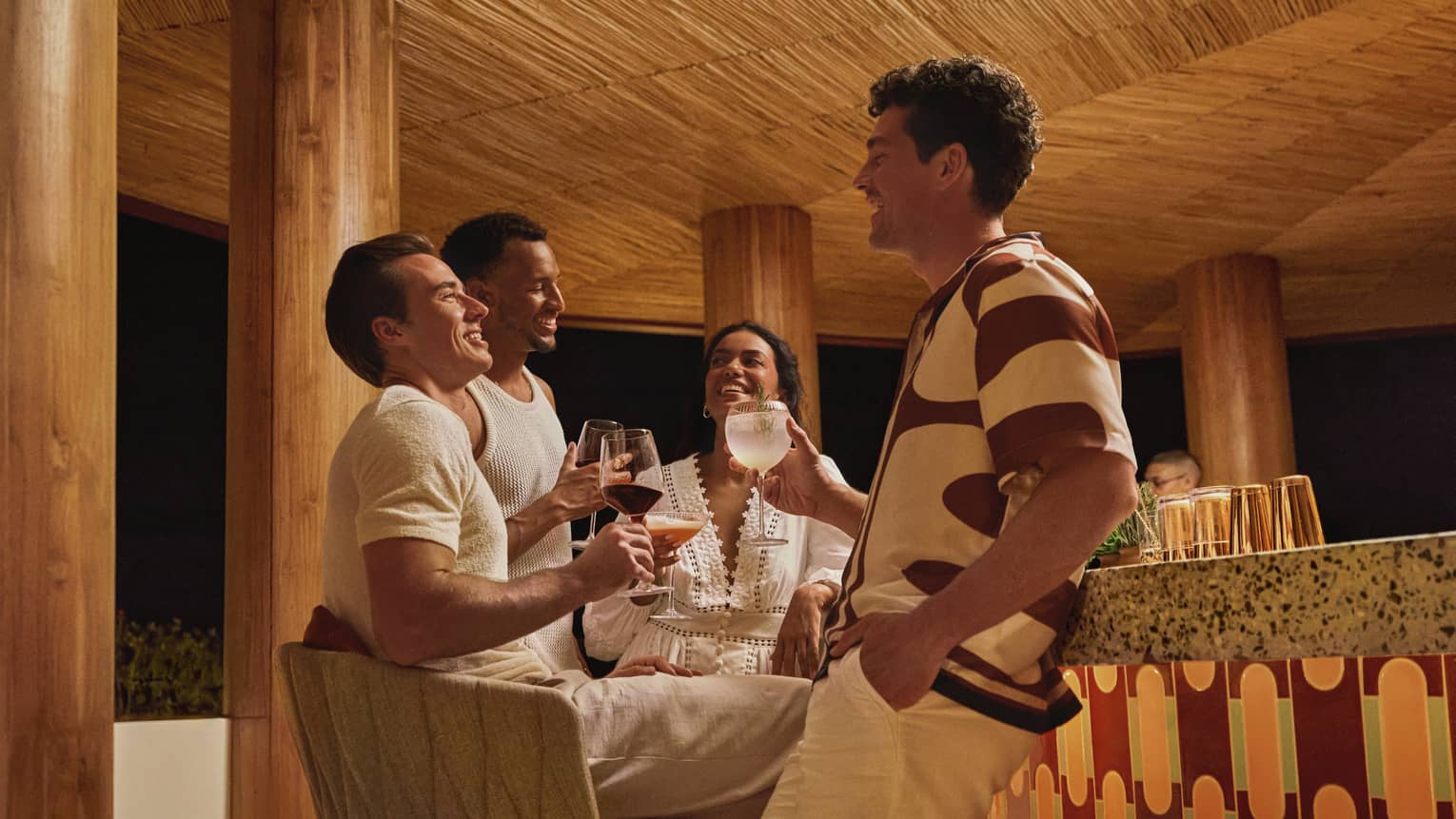 Four guests in stylish summer clothes hold cocktails and socialize next to a terazzo-topped bar in a round wooden pavilion.