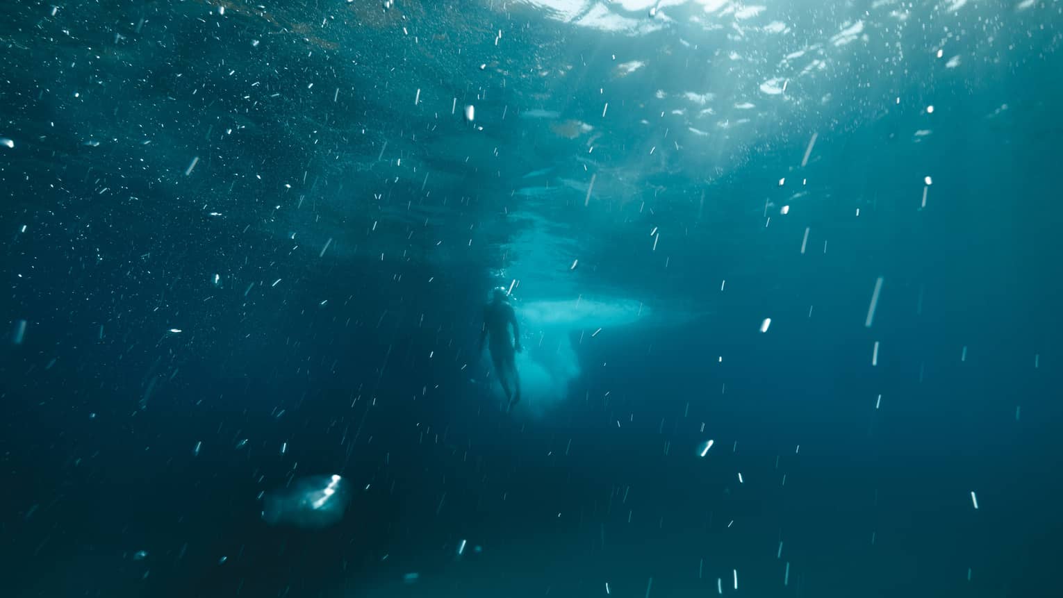 Deep underwater long view of a diver swimming toward the surface as sunlight pierces through the dark blue water.
