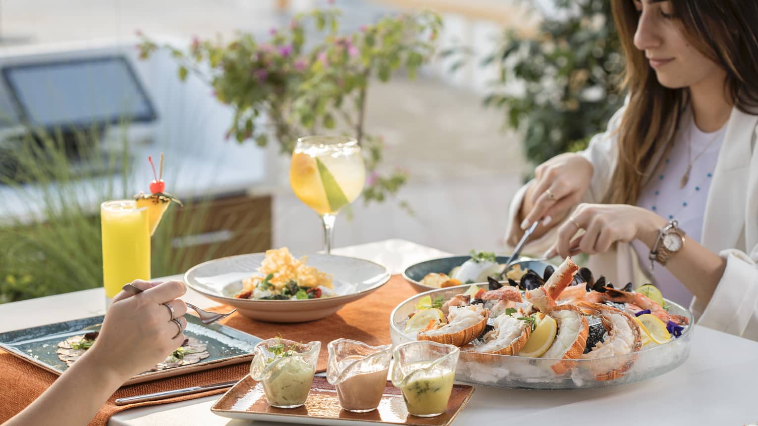 Two people sit at an outdoor table set with a large seafood platter on ice, a plate of three sauces and three other plates of food along with two tropical beverages