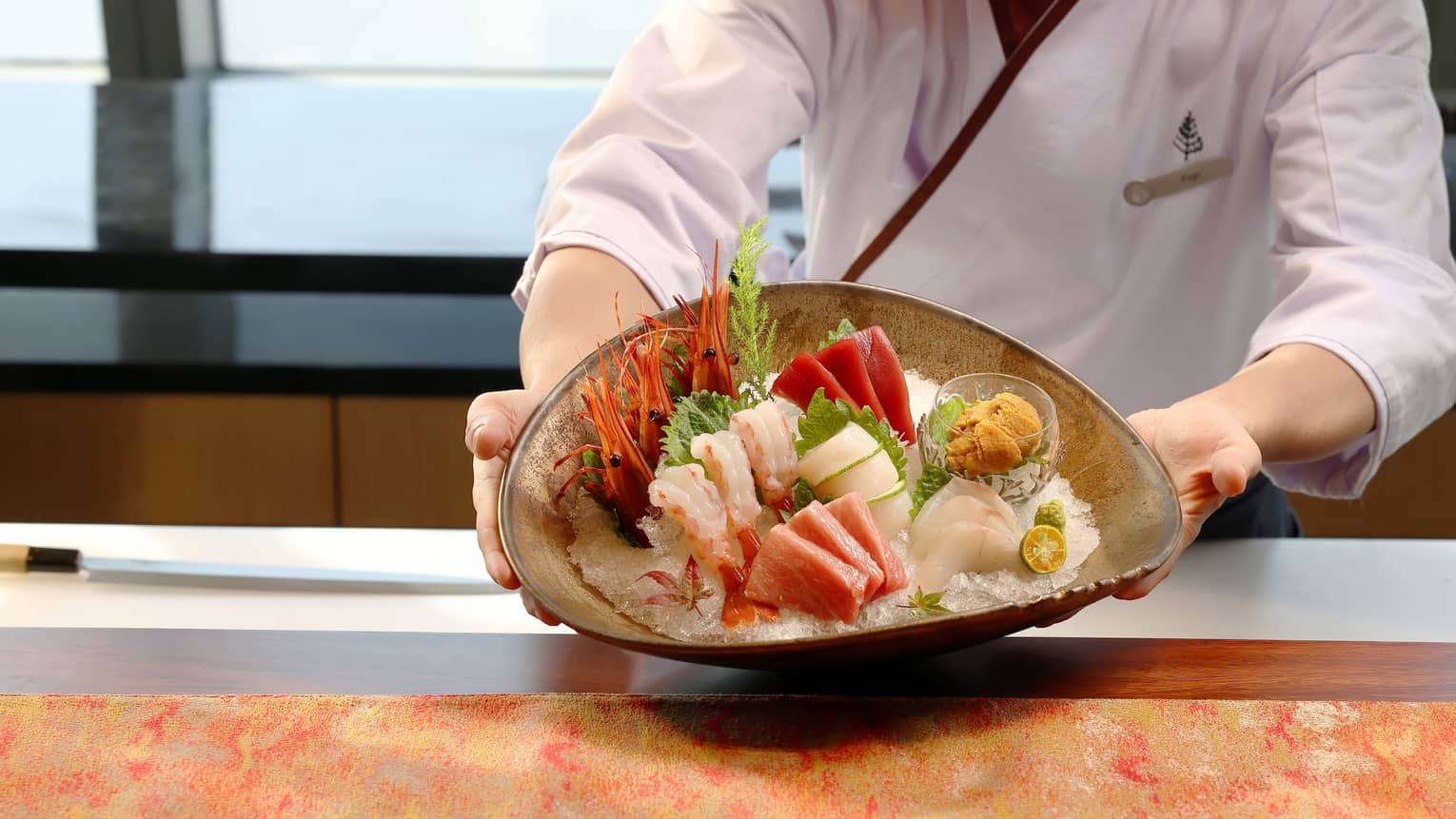 Close-up of a chef presenting a large bowl filled with vibrant prawns, shrimp, tuna and fresh garnishes on a bed of ice.