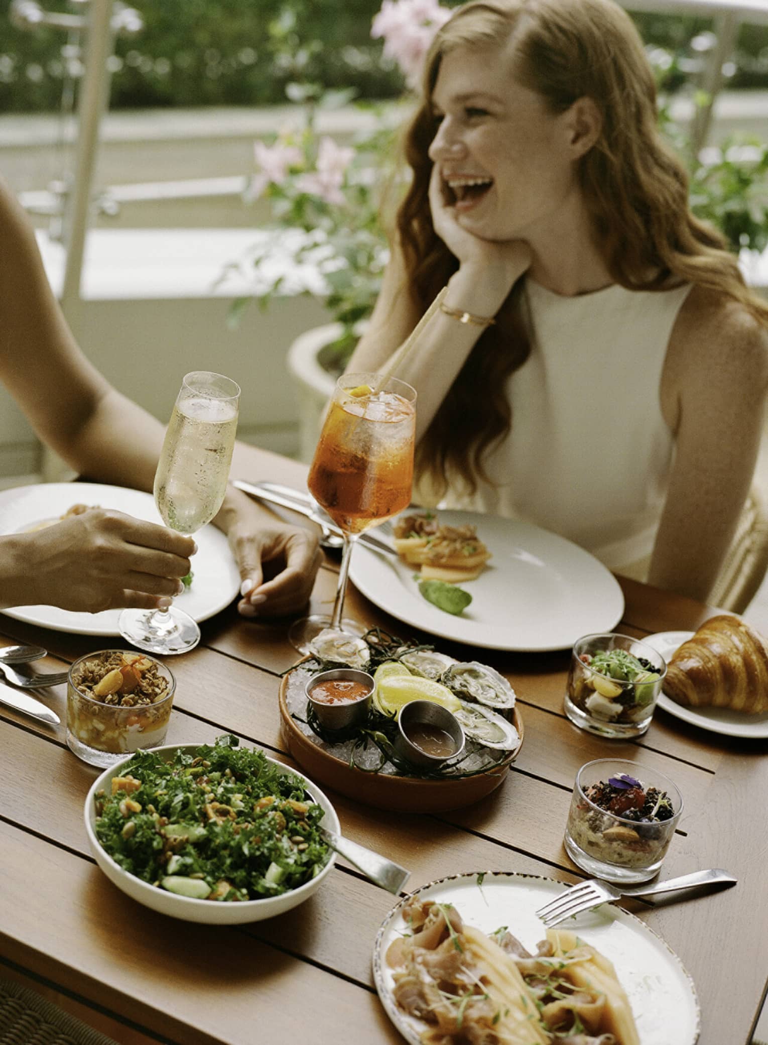 Laughing guests enjoying cold beverages, raw oysters with sauces, smoked salmon, a kale salad, muesli and croissants.