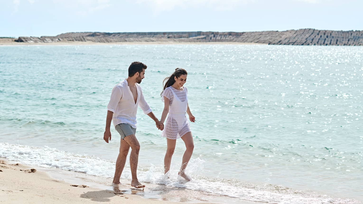 A man and woman both dressed in white hold hands and walk on a beach