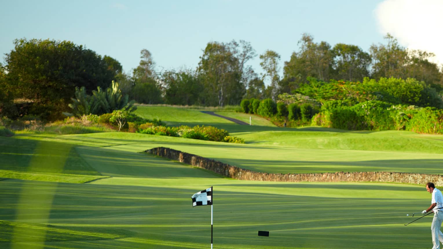 Man with golf club walks toward flag on golf course green near stone fence