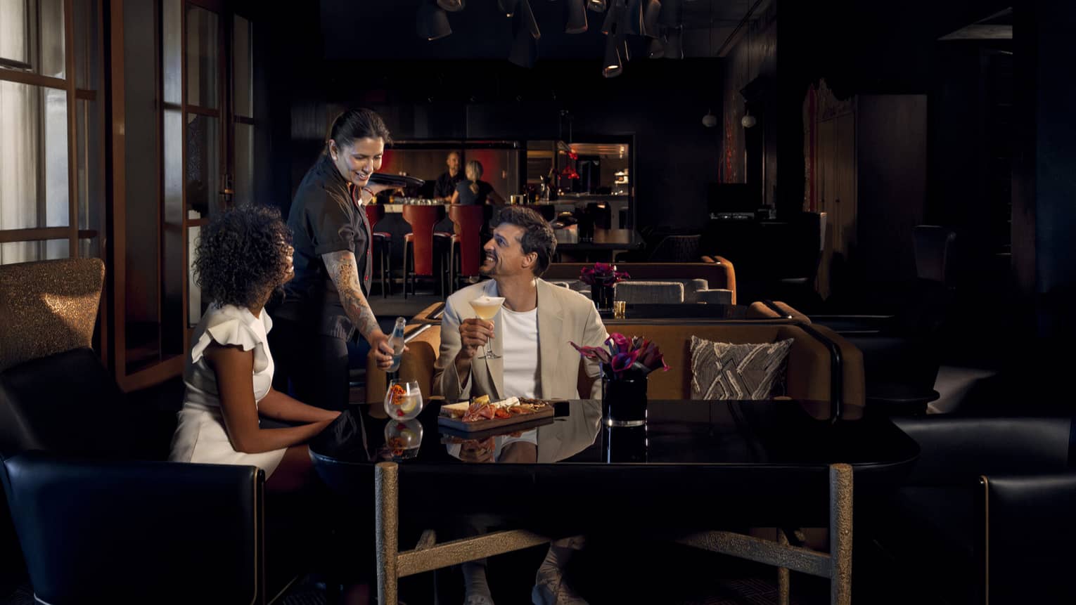 Two people wearing light-coloured cocktail attire sit on benches around a black table as a waitress brings them beverages