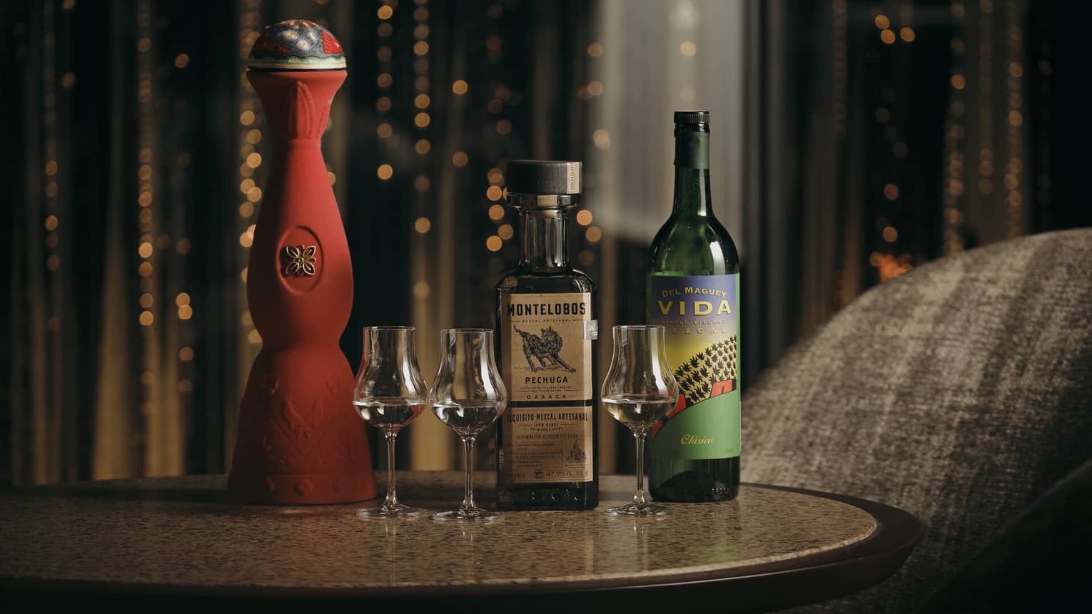 Tall, ornate red ceramic bottle alongside two more mezcal bottles and three tulip-shaped stemmed glasses of clear mezcal.