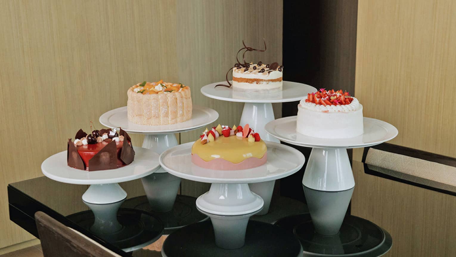 Five cakes of various colors and flavours, each on its own round, white cake stand are arranged together in a group on a black table