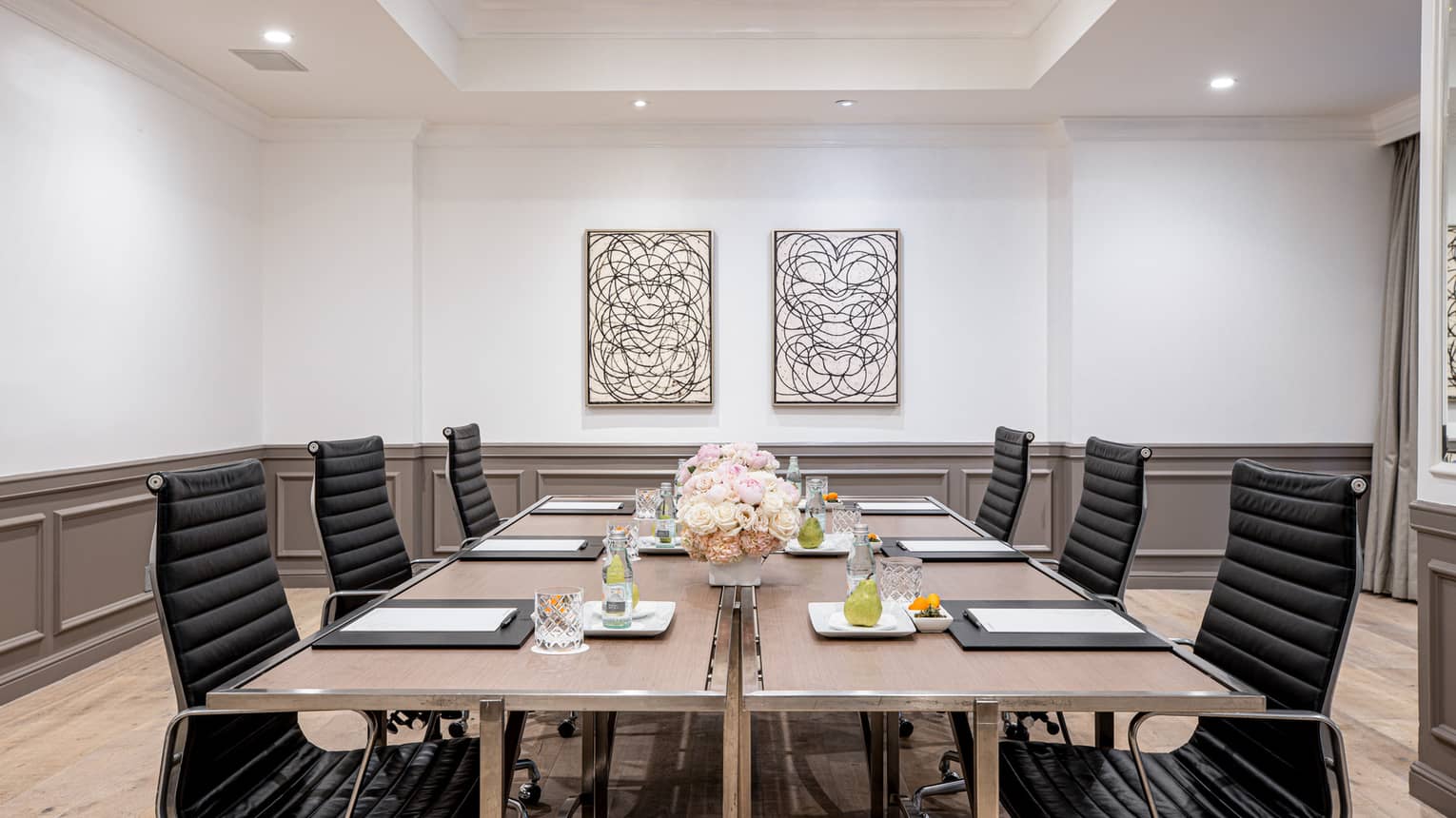 Six black chairs around a wooden table topped with writing pads, beverages and floral arrangements in a bright, spacious conference room