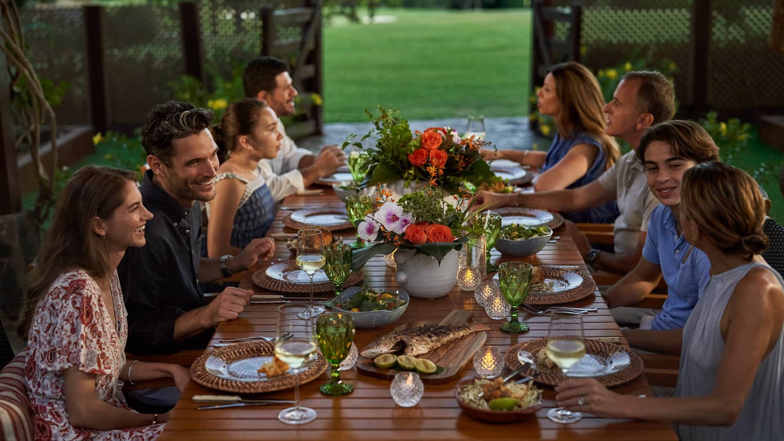 A large group sits at an outdoor dining table decorated with floral arrangements