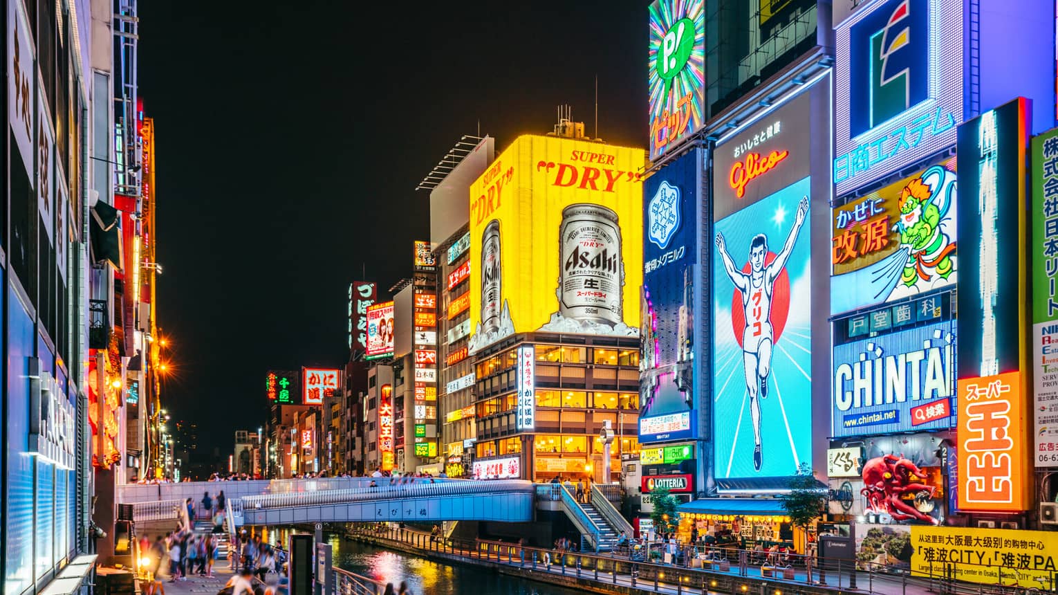 A footbridge crosses a narrow canal flanked by huge neon ads in bright colours on tall buildings in a busy city at night.