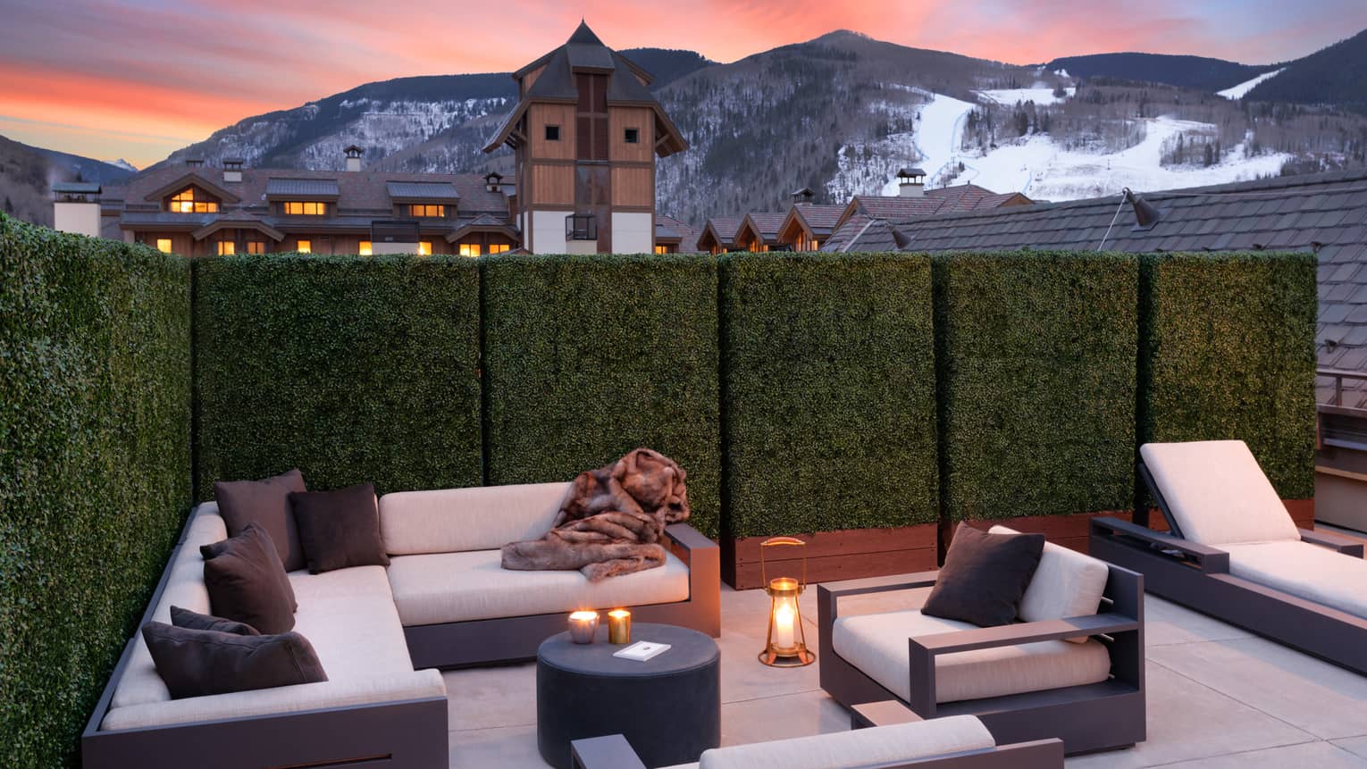 An outdoor seating area with. candles and white sofas near a cabin like village and mountains.