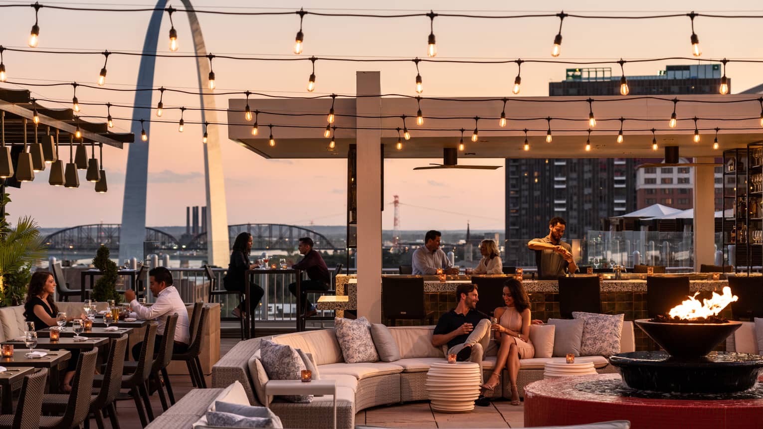 Cinderhouse outdoor patio with couples seated at banquette, sectional sofa, bar with pergola, view of arch at sunset