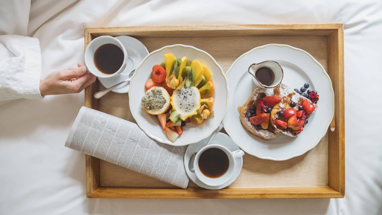 A wooden tray with various fruit, French toast and maple syrup served on white scalloped plates and two cups of coffee 