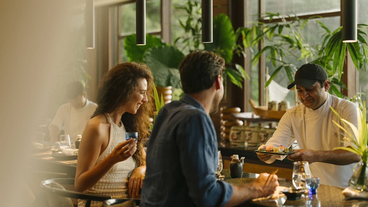 A couple is served sushi at a bar filed with natural light and lush green plants